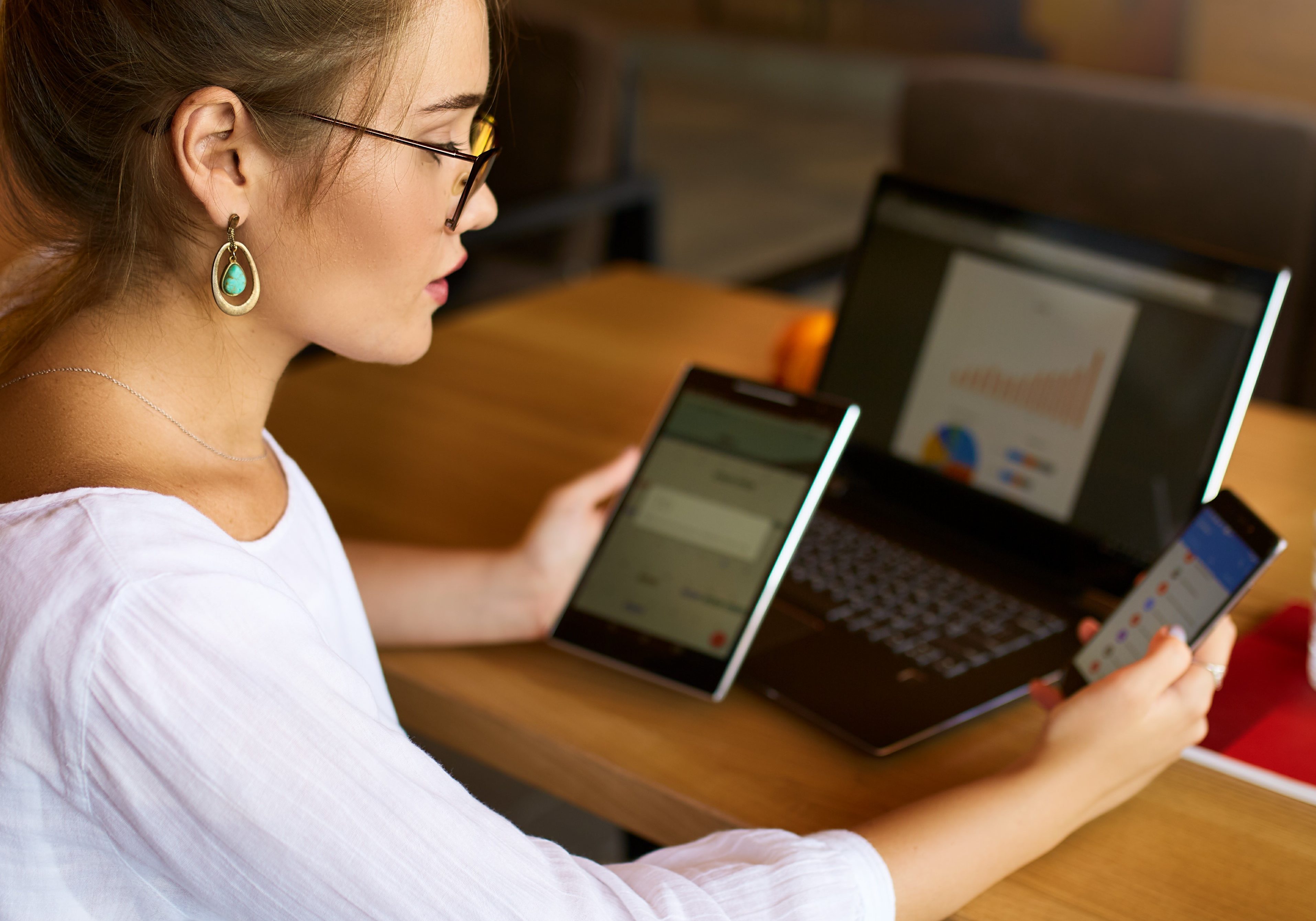 Mixed race woman in glasses working with multiple electronic internet devices. Freelancer businesswoman has tablet and smartphone in hands and laptop on table with charts on screen. Multitasking and telecommuting theme.